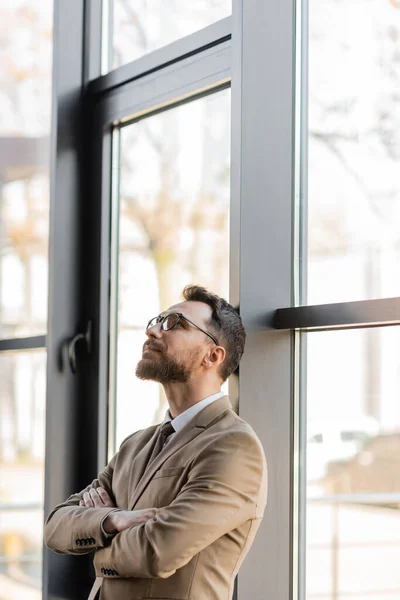 Fashionable and dreamy businessman in beige blazer and stylish eyeglasses standing with folded arms near large windows and looking up in modern office — Stock Photo