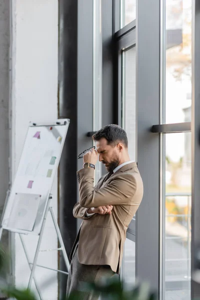 Side view of exhausted manager in trendy business attire holding eyeglasses and standing with closed eyes near large windows and flip chart with sticky notes in office — Stock Photo