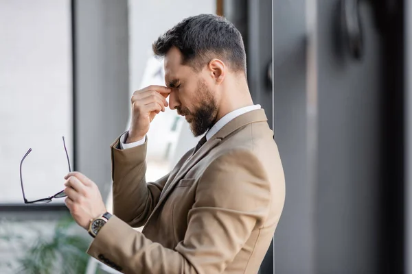 Side view of tired bearded businessman in beige trendy blazer and luxury wristwatch holding eyeglasses and touching nose bridge while standing with closed eyes in office — Stock Photo
