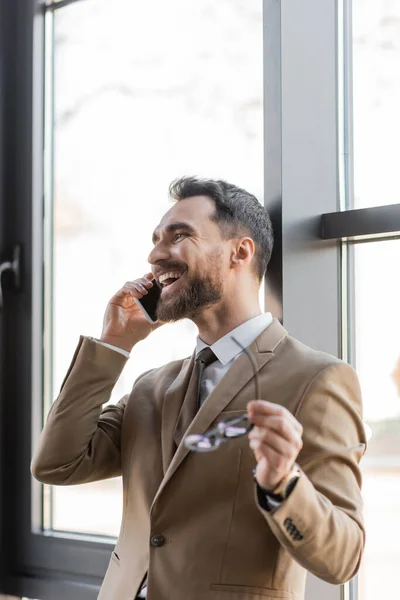 Imprenditore felice e di successo in giacca beige alla moda e cravatta parlando sul telefono cellulare e tenendo occhiali sfocati vicino alle finestre in ufficio moderno — Foto stock