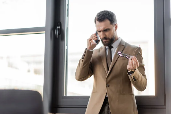 Стурбований бізнесмен в бежевому блістері і краватка тримає стильні окуляри і кидається під час розмови на мобільному телефоні біля великих вікон в сучасному офісі — стокове фото