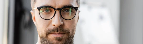 Retrato de bom aspecto e ambicioso empresário com barba e bigode vestindo óculos elegantes e olhando para a câmera no escritório borrado, banner — Fotografia de Stock