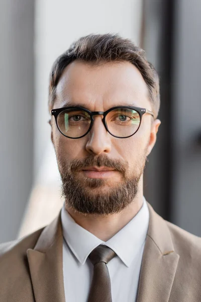 Портрет уверенного и харизматичного бизнесмена в стильной формальной одежде, такой как бежевый пиджак, галстук и очки, смотрящего на камеру в офисе на размытом фоне — стоковое фото