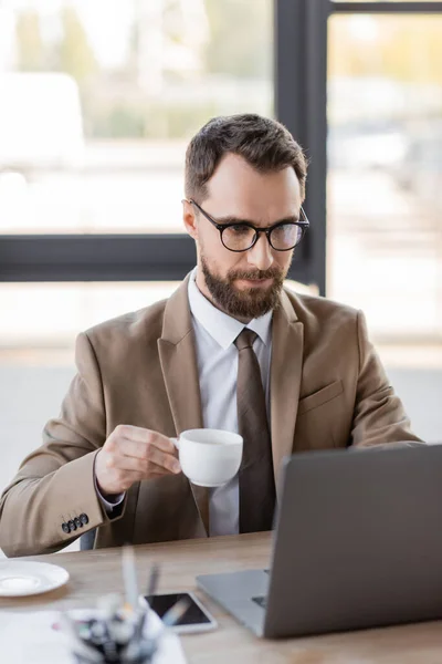 Mirato uomo d'affari barbuto in blazer elegante, occhiali e cravatta tenendo tazza di caffè vicino al telefono cellulare con schermo bianco e lavorando su laptop sfocato in ufficio — Foto stock