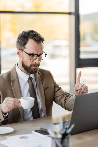Stringent bearded corporate manager in stylish business attire holding coffee cup and showing attention gesture during video conference on laptop near mobile phone and documents on work desk — Stock Photo