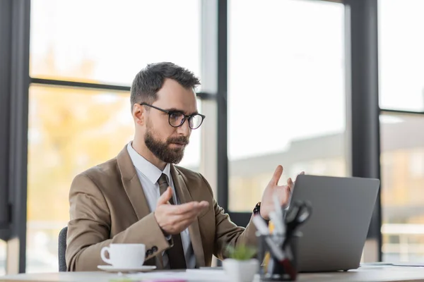Homem de negócios barbudo em óculos, blazer elegante e gesto gravata durante videoconferência no laptop perto de xícara de café e papelaria borrada no escritório — Fotografia de Stock