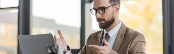 Entrepreneur barbu accompli et réussi en blazer beige, cravate et lunettes ayant chat vidéo sur ordinateur portable et geste dans le bureau moderne, bannière — Stock Photo