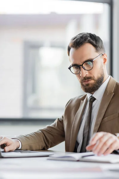 Manager corporatif concentré avec barbe et lunettes, vêtu d'un blazer et d'une cravate tendance, regardant un carnet flou et travaillant sur ordinateur au bureau — Photo de stock