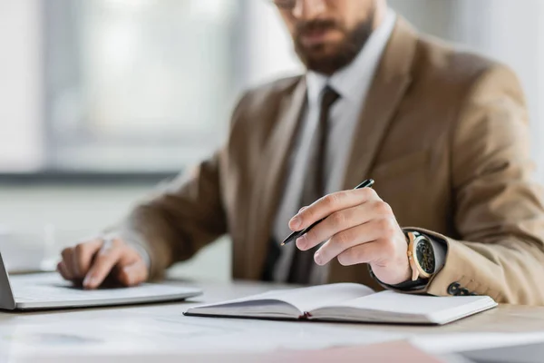 Partial view of corporate manager in beige blazer, tie and luxury wristwatch sitting with pen near notebook while working on laptop at workplace in office — Stock Photo