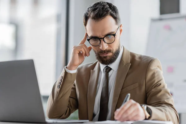 Nachdenkliche und konzentrierte bärtige Manager in trendiger Business-Kleidung berühren Kopf beim Schreiben der Planung in Notizbuch in der Nähe Laptop auf dem Schreibtisch im Büro — Stockfoto