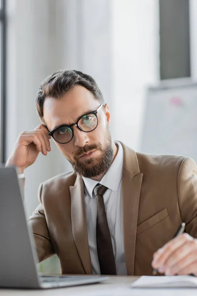 Serious bearded entrepreneur in tie, beige blazer and eyeglasses sitting with pen next to blurred laptop and notebook and looking at camera while holding hand near head — Stock Photo