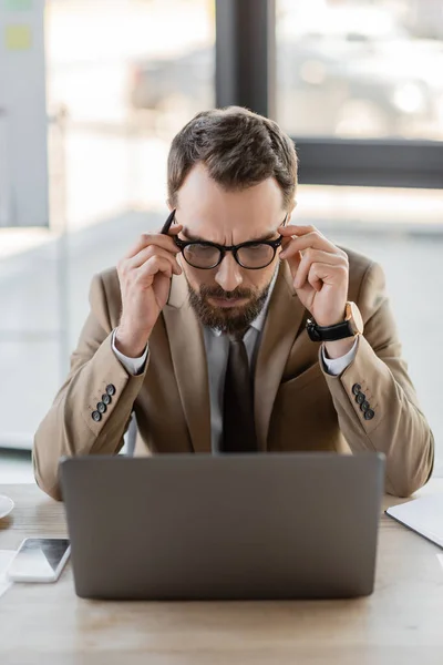 Empresário exausto em blazer bege e gravata tocando óculos enquanto sofre de dor de cabeça perto de laptop e telefone celular com tela em branco no escritório — Fotografia de Stock