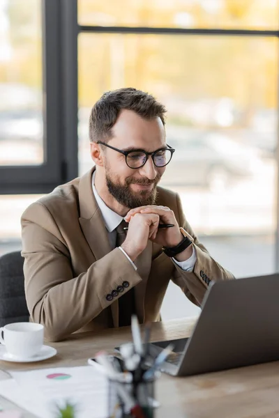 Empresário satisfeito e carismático em óculos e blazer bege sorrindo durante o bate-papo por vídeo no laptop enquanto sentado na mesa de trabalho perto de xícara de café e documentos no escritório — Fotografia de Stock