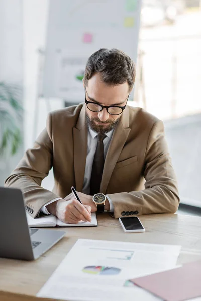 Successful entrepreneur in trendy blazer, eyeglasses and luxury wristwatch writing in notebook next to laptop, smartphone with blank screen and documents with charts on desk in office — Stock Photo