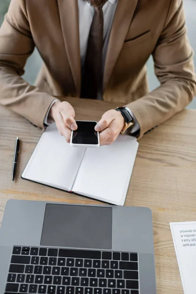 Vista parcial del hombre de negocios en blazer beige que sostiene el teléfono inteligente con la pantalla en blanco cerca del cuaderno vacío, el ordenador portátil, la pluma y el documento en el escritorio en la oficina, vista de ángulo alto - foto de stock