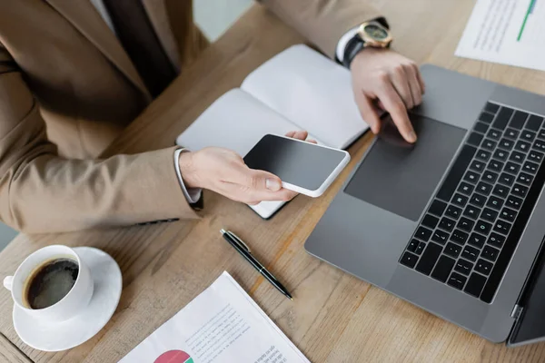 Partial view of stylish entrepreneur holding mobile phone with blank screen while using touchpad of laptop near empty notebook, pen, coffee cup and documents at workplace in office, high angle view — Stock Photo