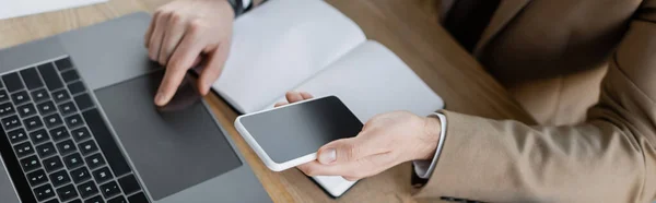 High angle view of cropped businessman in beige formal wear working on computer and holding cellphone with blank screen near empty notepad on office desk, banner — Stock Photo