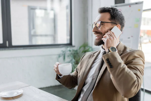 Empresário alegre e bem sucedido em blazer elegante bege, óculos e relógio de pulso sentado com xícara de café perto de pires na mesa de escritório e falando no telefone móvel — Fotografia de Stock