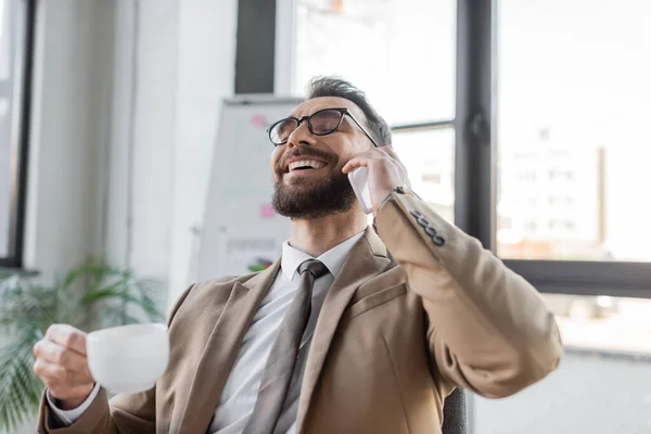 Overjoyed and accomplished entrepreneur in trendy business attire holding coffee cup and talking on mobile phone near flip chart of blurred background in office — Stock Photo