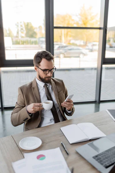 High angle view of manager in beige blazer, tie and eyeglasses holding coffee cup and looking at laptop while sitting at workplace near empty notebook, documents and saucer on office desk, blur — Stock Photo