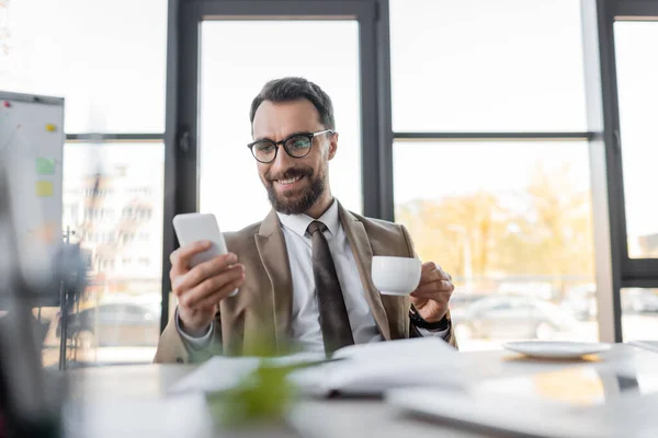 Ambitious corporate manager in beige stylish blazer, eyeglasses and tie holding coffee cup and looking at mobile phone while smiling near notebook, saucer and computer on blurred foreground — Stock Photo