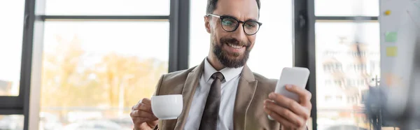 Homme d'affaires barbu joyeux dans blazer branché, cravate et lunettes tenant tasse de café et regardant le téléphone mobile sur le premier plan flou dans le bureau, bannière — Photo de stock