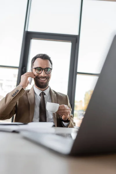 Hombre de negocios carismático con barba y cabello moreno, vistiendo chaqueta y corbata beige, hablando en el teléfono inteligente y sosteniendo la taza de café frente a la computadora portátil en primer plano borroso en la oficina - foto de stock