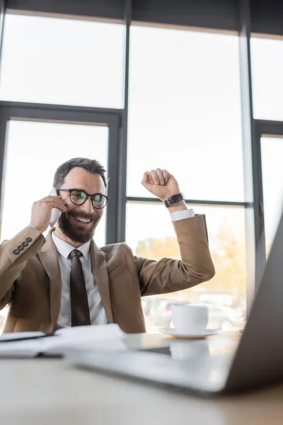 Overjoyed bearded manager in stylish business attire talking on mobile phone and showing win gesture near cup of coffee and computer on blurred foreground in office — Stock Photo