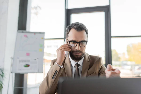 Serious bearded businessman in eyeglasses, beige blazer and tie talking on mobile phone and pointing at laptop near flip chart on blurred background in modern office — Stock Photo