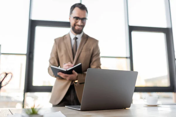 Homem de negócios barbudo positivo em óculos, blazer bege e gravata de pé perto do laptop e xícara de café na mesa de trabalho e escrevendo em notebook sobre fundo borrado — Fotografia de Stock