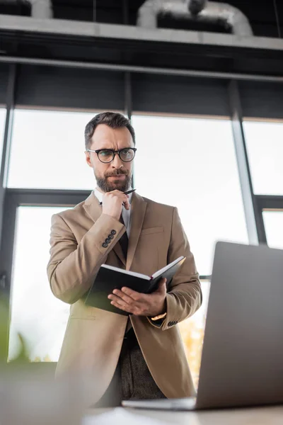 Low angle view of thoughtful businessman in eyeglasses and stylish blazer standing with notebook and pen and looking at laptop in office on blurred foreground — Stock Photo