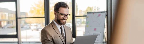 Веселый бородатый менеджер в очках, бежевый пиджак и галстук глядя на ноутбук во время видеоконференции возле флип-чарта с графиками и аналитикой в офисе, баннер — стоковое фото