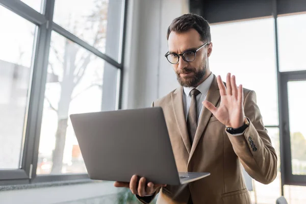 Serious bearded businessman in trendy eyeglasses, beige blazer and tie holding laptop and waving hand while having video conference near blurred windows in office — Stock Photo