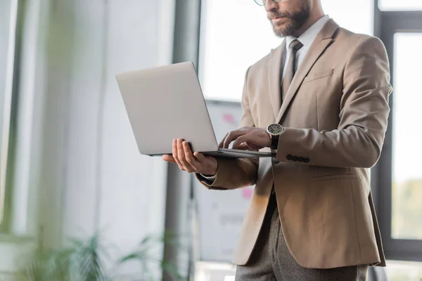 Ausgeschnittene Ansicht eines vollbärtigen Unternehmers in beigem Blazer, Krawatte und Luxus-Armbanduhr mit Laptop im Büro auf verschwommenem Hintergrund — Stockfoto