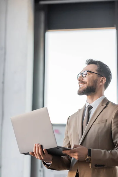 Happy and satisfied corporate manager in eyeglasses and fashionable business attire such as beige blazer and tie standing with laptop and looking away in office — Stock Photo
