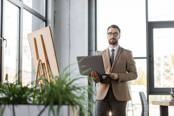 Ambicioso empresário barbudo em óculos, blazer bege e gravata de pé com laptop e olhando para a câmera perto de flip chart, grandes janelas e plantas borradas no escritório — Fotografia de Stock