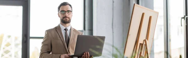 Confident professional entrepreneur in beige blazer, eyeglasses and tie holding laptop and looking at camera while standing near flip chart in contemporary office, banner — Stock Photo