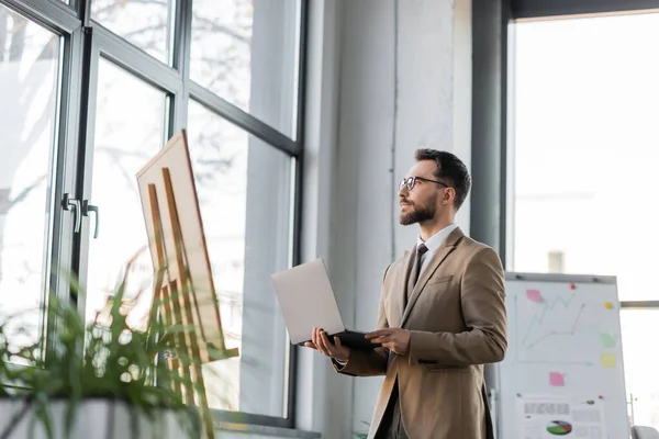 Confident and fashionable bearded businessman with laptop looking away through windows near flip charts with infographics and blurred plants in contemporary office — Stock Photo