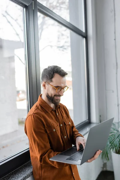 Empreendedor barbudo positivo e bem sucedido em óculos da moda e camisa elegante de pé perto de grandes janelas no escritório contemporâneo e trabalhando no laptop — Fotografia de Stock