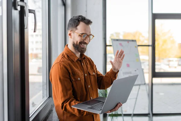 Good looking and positive bearded businessman in eyeglasses and trendy shirt smiling and waving hand during video call on laptop near flip chart on blurred background in office — Stock Photo
