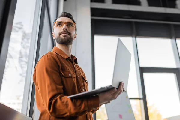 Low angle view of thoughtful bearded businessman in trendy shirt and stylish eyeglasses holding laptop while standing in modern office and looking away, corporate lifestyle — Stock Photo