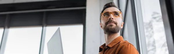 Low angle view of charismatic and brunette businessman with beard, wearing fashionable eyeglasses and shirt, looking away and thinking in office, banner, corporate lifestyle — Stock Photo
