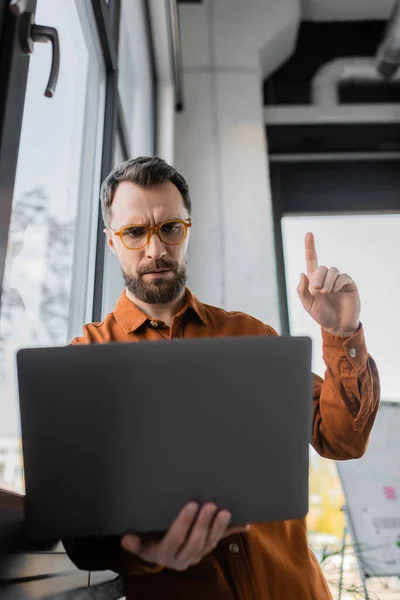 Worried bearded businessman in stylish eyeglasses and shirt pointing up with finger while showing attention gesture during video conference on laptop in office, corporate lifestyle — Stock Photo