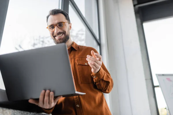 Low angle view of pleased bearded entrepreneur in fashionable eyeglasses and shirt gesturing during video conference on laptop near window in modern office, corporate lifestyle — Stock Photo