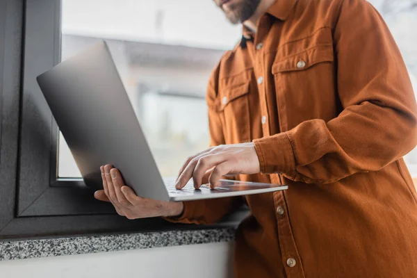 Cropped view of successful corporate manager in fashionable casual shirt standing near window in contemporary office and using laptop on blurred background, corporate lifestyle — Stock Photo
