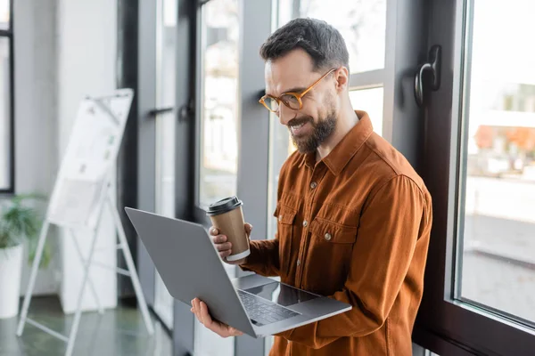 Brunette and bearded businessman in eyeglasses and stylish shirt holding disposable cup with coffee and looking at laptop near flip chart on blurred background in office — Stock Photo