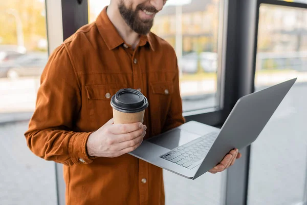 Partial view of smiling corporate manager in stylish shirt standing with laptop and takeaway drink in paper cup near windows in modern office on blurred background — Stock Photo