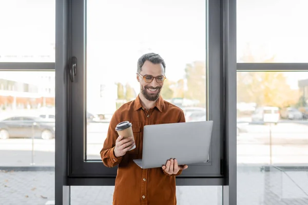 Pleased and bearded businessman in eyeglasses and stylish shirt smiling while standing with takeaway drink and laptop near windows in contemporary office — Stock Photo