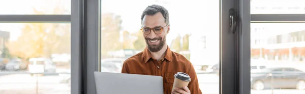 Задоволений бородатий бізнесмен в модних окулярах і сорочках тримає ноутбук і паперовий стаканчик з виносним напоєм, стоячи біля вікон в офісі, банер, корпоративний спосіб життя — стокове фото
