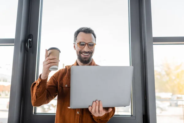 Successful bearded businessman in trendy shirt and eyeglasses holding coffee to go and pointing up with finger during video call on laptop in modern office, corporate lifestyle — Stock Photo